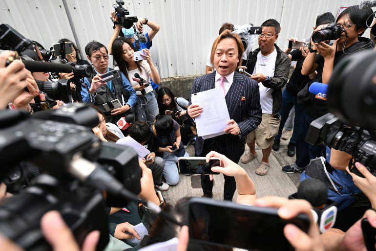 Lawrence Lau speaks to the media.  He wears a striped suit and holds a document.  Journalists are all around him.  Some are equipped with cameras.