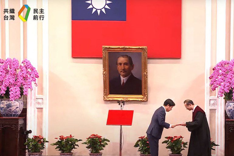 William Lai Ching-ti during his inauguration.  There is a Taiwan flag on the wall and a portrait of Sun Yat-sen, the founder of the Republic of China.  With large displays of pink orchids on either side.  Lai bends down as he receives a document from an official.